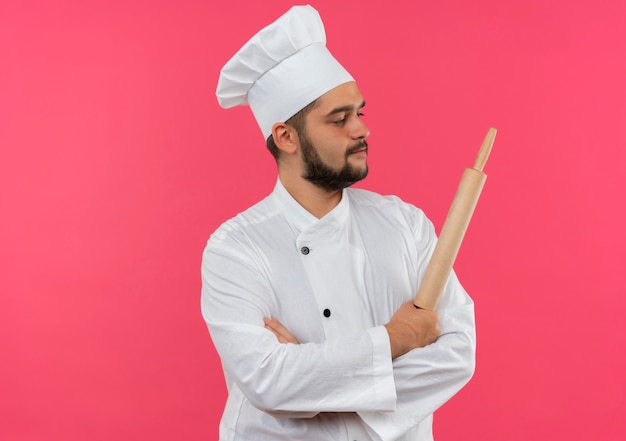 Jeune cuisinier confiant en uniforme de chef debout avec une posture fermée et tenant un rouleau à pâtisserie regardant de côté isolé sur un mur rose avec espace de copie