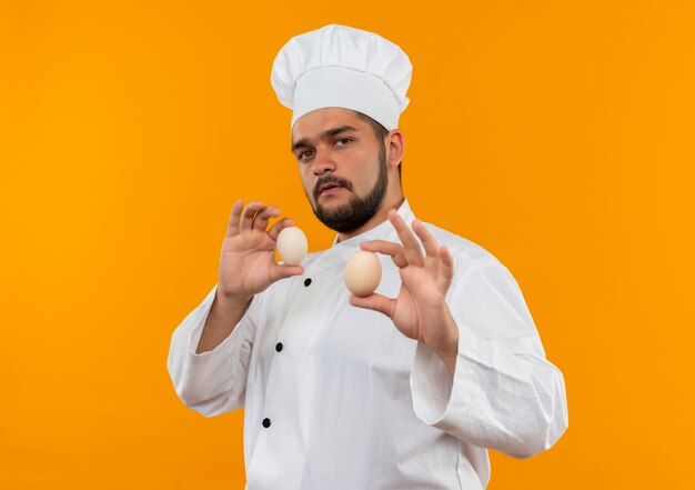 Jeune cuisinier confiant en uniforme de chef tenant des œufs isolés sur un mur orange