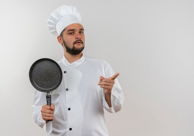 Jeune cuisinier confiant en uniforme de chef tenant une poêle à frire et pointant tout droit isolé sur un mur blanc avec espace de copie