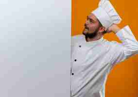 Photo gratuite jeune cuisinier confus en uniforme de chef debout derrière et regardant un mur blanc avec la main derrière la tête isolée sur un mur orange avec espace de copie