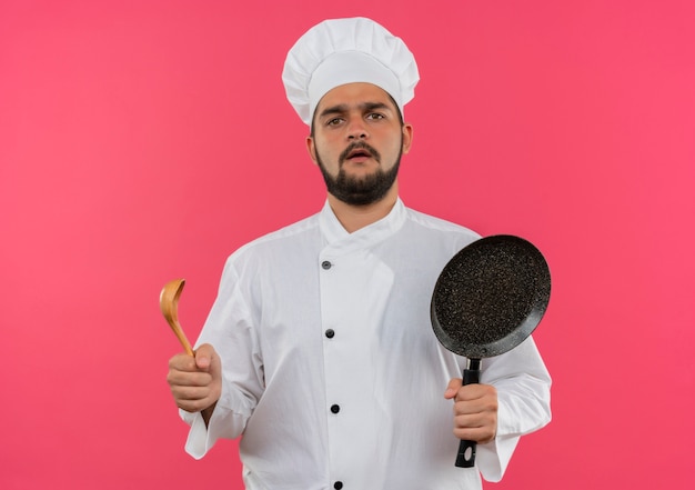 Jeune cuisinier confus en uniforme de chef tenant une poêle à frire et une cuillère isolée sur un mur rose