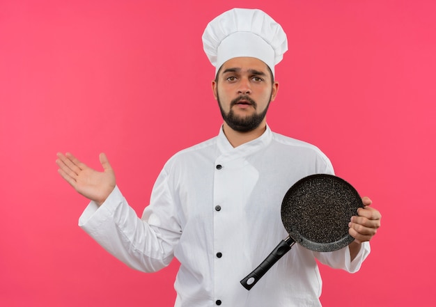 Jeune cuisinier confus en uniforme de chef tenant une poêle à frire et montrant une main vide isolée sur un mur rose