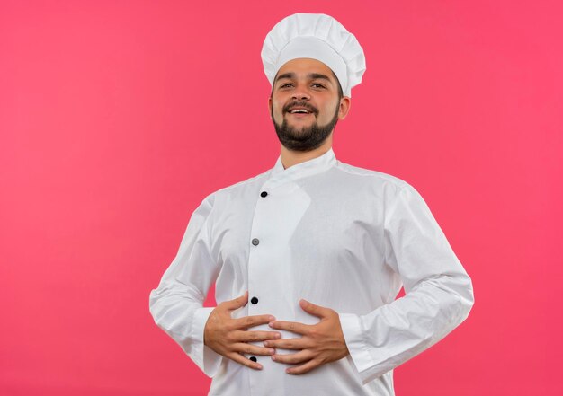 Jeune cuisinier joyeux en uniforme de chef mettant les mains sur le ventre isolé sur un mur rose avec espace de copie