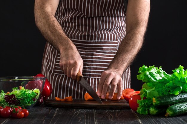 Jeune cuisinier à la maison homme en tablier trancher la carotte avec un couteau de cuisine. Les mains des hommes coupent la carotte pour faire une salade sur fond noir. Concept d'aliments sains
