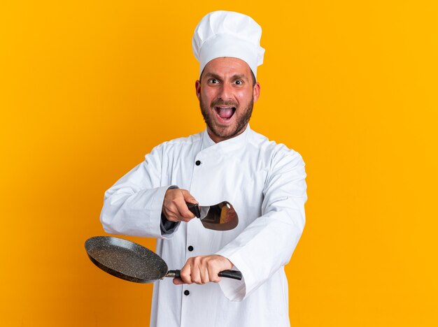 Jeune cuisinier masculin caucasien furieux en uniforme de chef et casquette regardant la caméra tenant une poêle à frire pointant vers la caméra avec un couperet criant isolé sur un mur orange avec un espace de copie