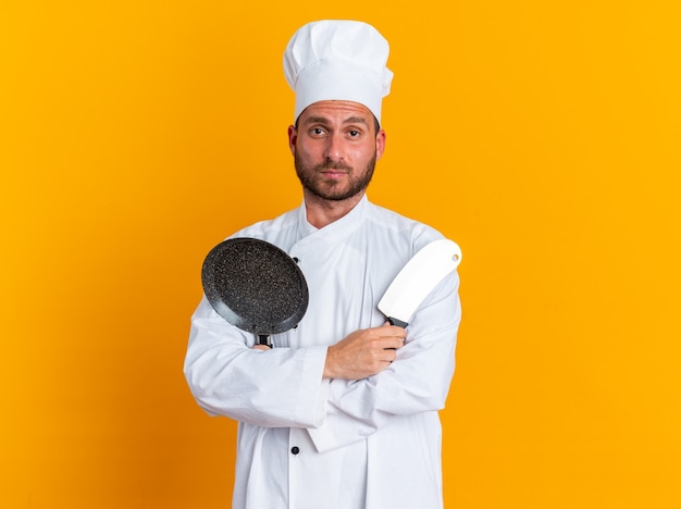 Jeune cuisinier masculin caucasien sérieux en uniforme de chef et casquette debout avec une posture fermée tenant un couperet et une poêle à frire regardant la caméra isolée sur un mur orange avec espace de copie