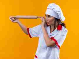 Photo gratuite jeune cuisinière concernée portant un uniforme de chef tenant et regardant un rouleau à pâtisserie isolé sur un mur orange