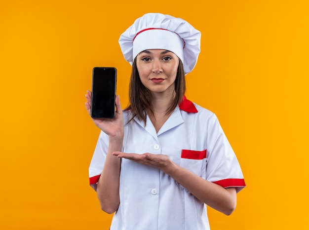 jeune cuisinière confiante portant l'uniforme du chef tenant et pointe avec la main au téléphone isolé sur un mur orange