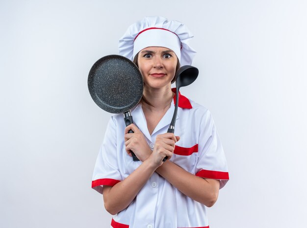 Jeune cuisinière confuse portant un uniforme de chef tenant et traversant une poêle à frire avec une louche isolée sur fond blanc