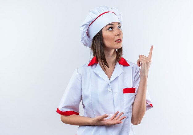 Jeune cuisinière impressionnée portant des points uniformes de chef vers le haut isolé sur un mur blanc