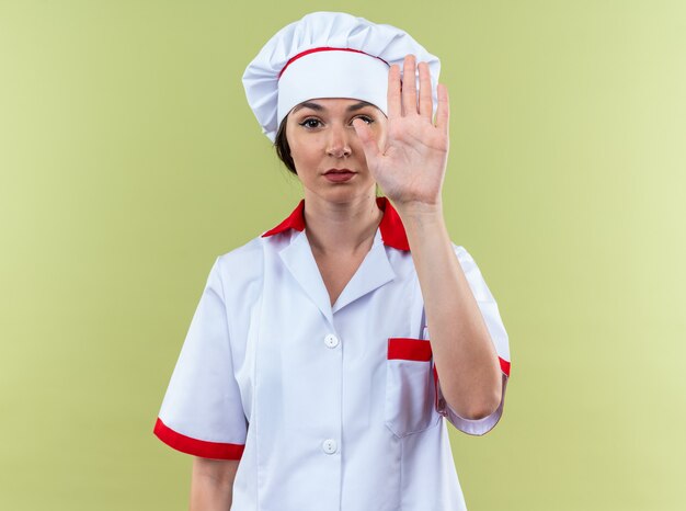 Jeune cuisinière stricte portant un uniforme de chef montrant un geste d'arrêt isolé sur un mur vert olive