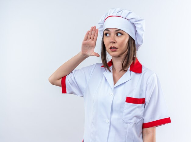 Jeune cuisinière suspecte portant l'uniforme du chef montrant le geste d'écoute isolé sur fond blanc