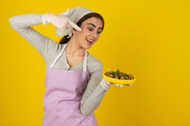 Jeune cuisinière en tablier tenant une assiette de champignons frits sur un mur jaune.