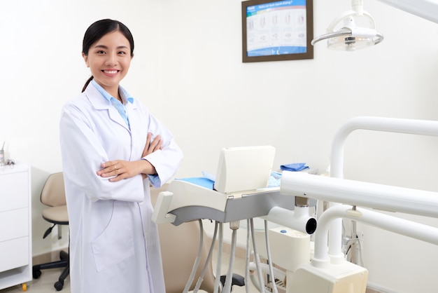 Jeune dentiste asiatique en blouse blanche qui pose en clinique à côté de l'équipement