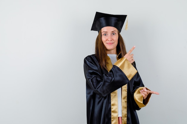 Photo gratuite jeune diplômée pointant de côté en tenue académique et à la recherche de confiance. vue de face.