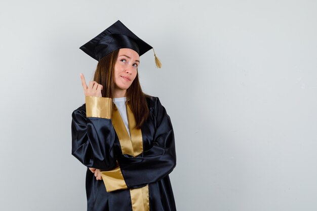 Jeune diplômée pointant vers le haut en tenue académique et à la recherche de confiance. vue de face.