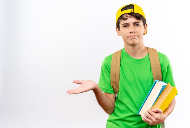 Jeune écolier confus portant un sac à dos avec une casquette tenant des livres écartant la main