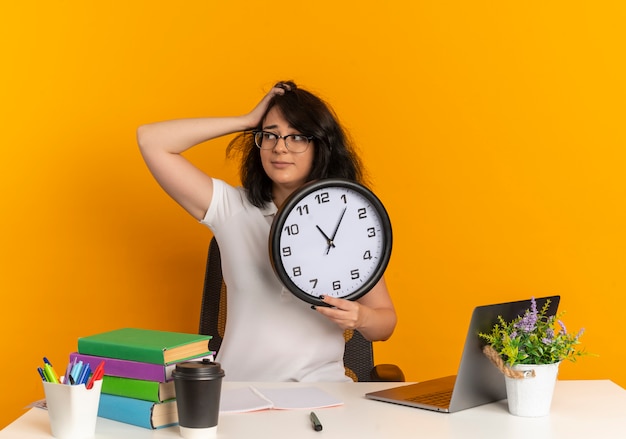 Jeune écolière assez caucasienne anxieuse portant des lunettes est assise au bureau avec des outils scolaires met la main sur la tête et tient l'horloge isolée sur l'espace orange avec copie espace