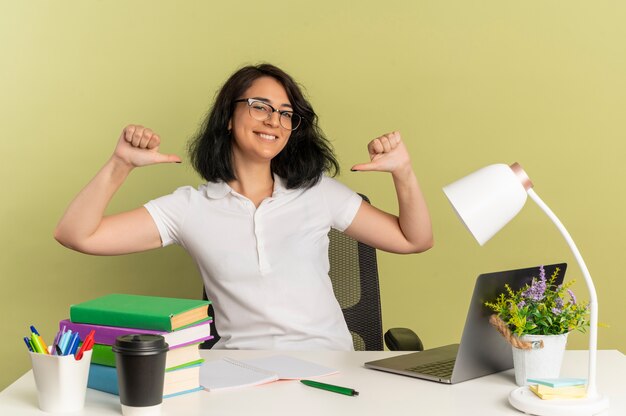 Jeune écolière assez caucasienne souriante portant des lunettes est assise au bureau avec des outils scolaires points à elle-même isolé sur un espace vert avec espace copie