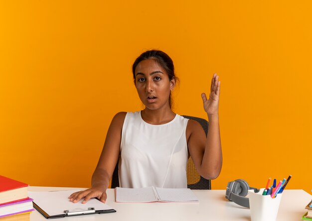 jeune écolière assis au bureau avec des outils scolaires levant la main