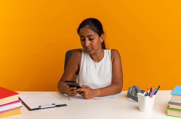 Jeune écolière assise au bureau avec des outils scolaires tenant et regardant le téléphone isolé sur le mur orange