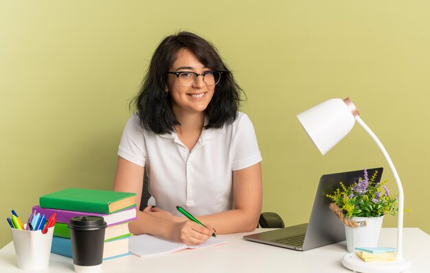 Jeune écolière caucasienne jolie souriante portant des lunettes est assise au bureau avec des outils scolaires tient un stylo isolé sur un espace vert avec espace copie