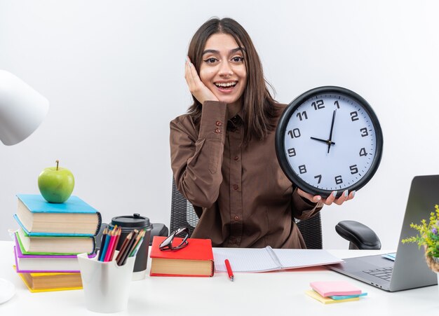 Une jeune écolière excitée est assise à table avec des outils scolaires tenant une horloge murale mettant la main sur la joue