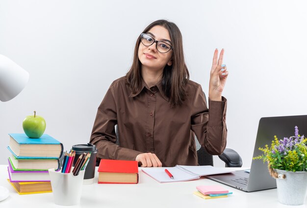 Une jeune écolière heureuse portant des lunettes est assise à table avec des outils scolaires montrant un geste de paix