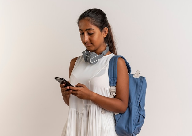 Jeune écolière portant un sac à dos et un casque à composer le numéro sur le téléphone sur blanc
