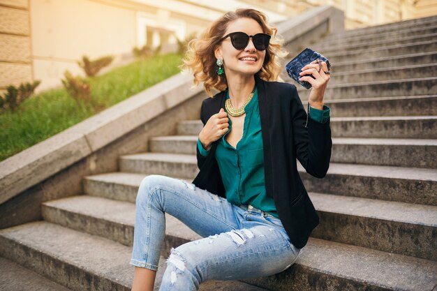 Jeune élégante belle femme assise sur l'escalier dans la rue de la ville, portant des jeans, veste noire, chemisier vert, lunettes de soleil, tenant sac à main, style élégant, tendance de la mode estivale, souriant