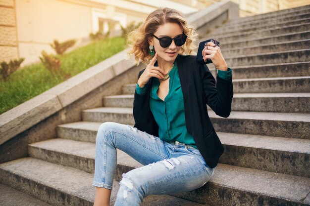 Jeune élégante belle femme assise sur l'escalier dans la rue de la ville, portant des jeans, veste noire, chemisier vert, lunettes de soleil, tenant sac à main, style élégant, tendance de la mode estivale, souriant