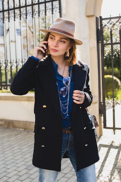 Jeune élégante jolie femme souriante et parler sur son téléphone, vêtue d'un manteau bleu foncé et d'un jean