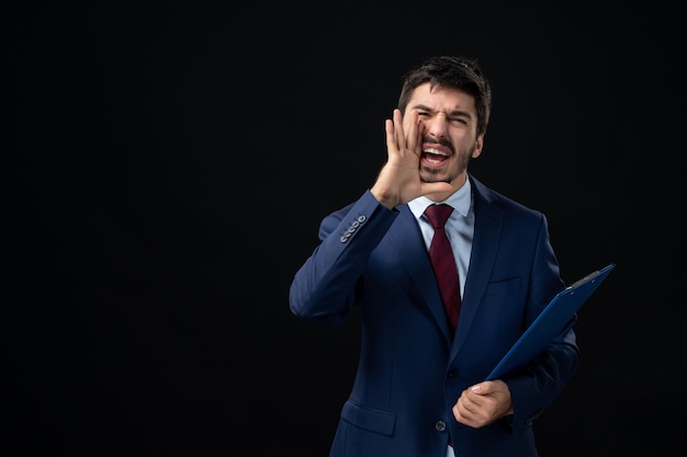 Jeune employé de bureau masculin en costume tenant des documents et appelant quelqu'un sur un mur sombre isolé