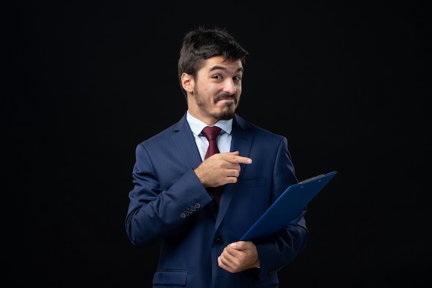 Photo gratuite jeune employé de bureau masculin souriant en costume tenant et pointant des documents sur un mur sombre isolé