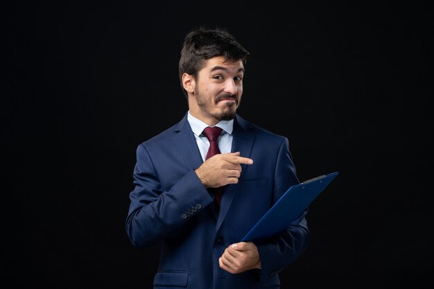 Jeune employé de bureau masculin souriant en costume tenant et pointant des documents sur un mur sombre isolé