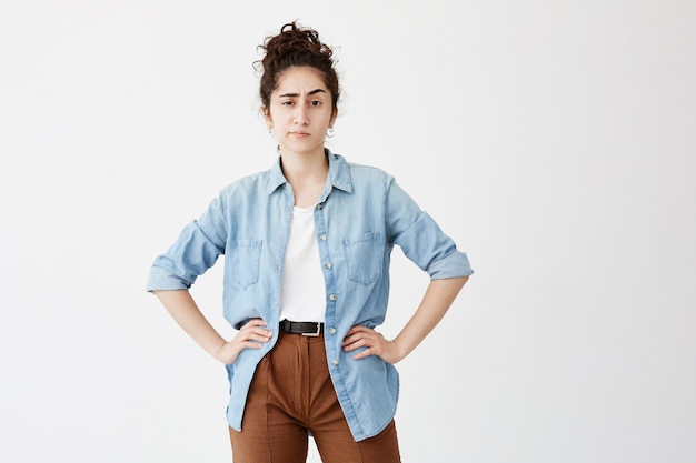 Jeune employée se reposant à la maison après le travail. Jolie étudiante aux cheveux bouclés noirs en chignon, habillée avec désinvolture en attendant ses amis, debout avec les mains sur les hanches contre le mur blanc