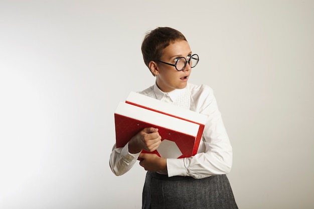 Jeune enseignant européen attrayant en chemisier blanc et jupe grise tenant des liants rouges et blancs roule les yeux d'incrédulité sur le mur blanc