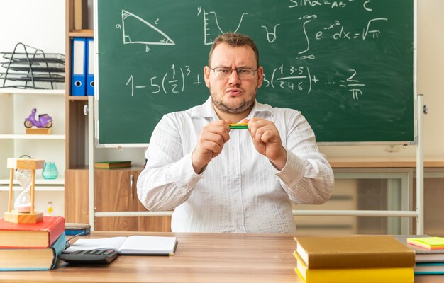 jeune enseignant fronçant les sourcils portant des lunettes assis au bureau avec des fournitures scolaires en classe tenant des bâtons de comptage horizontalement essayant de les casser en regardant à l'avant