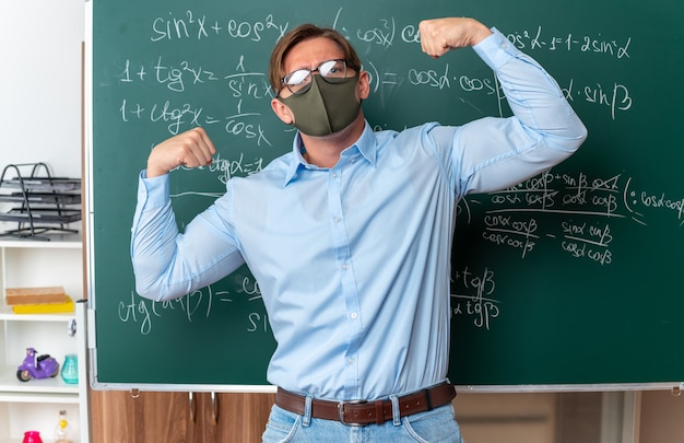 Jeune enseignant portant des lunettes dans un masque de protection faciale levant les poings comme un gagnant à l'air confiant debout près du tableau noir avec des formules mathématiques en classe