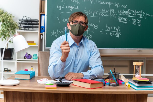 Jeune enseignant portant des lunettes et un masque de protection faciale montrant un crayon avec un visage sérieux assis au bureau de l'école avec des livres et des notes devant le tableau noir en classe