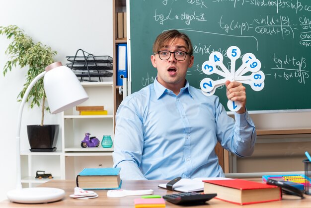 Jeune enseignant portant des lunettes montrant des plaques d'immatriculation expliquant la leçon à la surprise d'être assis au bureau de l'école avec des livres et des notes devant le tableau noir en classe