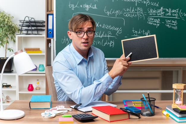 Photo gratuite jeune enseignant portant des lunettes avec un petit tableau noir et un morceau de craie expliquant la leçon à l'air confiant assis au bureau de l'école avec des livres et des notes devant le tableau noir en classe