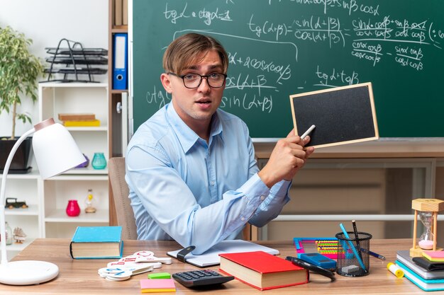 Jeune enseignant portant des lunettes avec un petit tableau noir et un morceau de craie expliquant la leçon à l'air confiant assis au bureau de l'école avec des livres et des notes devant le tableau noir en classe