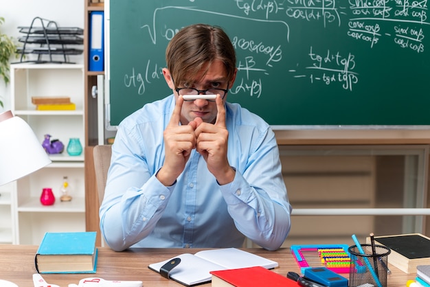 Jeune enseignant portant des lunettes tenant de la craie regardant avec un visage sérieux assis au bureau de l'école avec des livres et des notes devant le tableau noir en classe