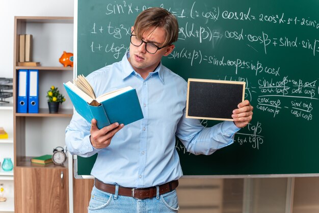 Jeune enseignant portant des lunettes tenant un petit livre de lecture de tableau noir à la confiance debout près du tableau noir avec des formules mathématiques en classe