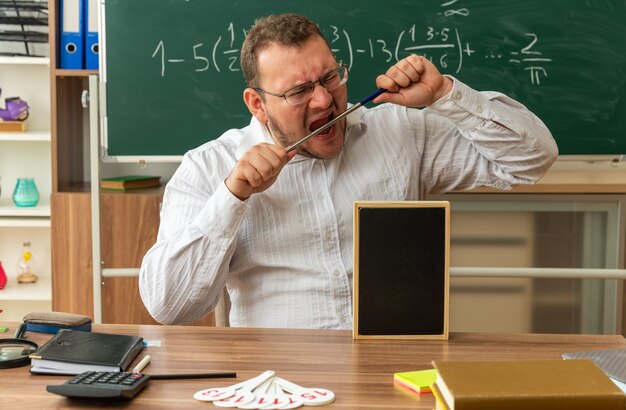 jeune enseignant stressé portant des lunettes assis au bureau avec des fournitures scolaires et un mini tableau noir dessus en classe mordant le bâton de pointeur