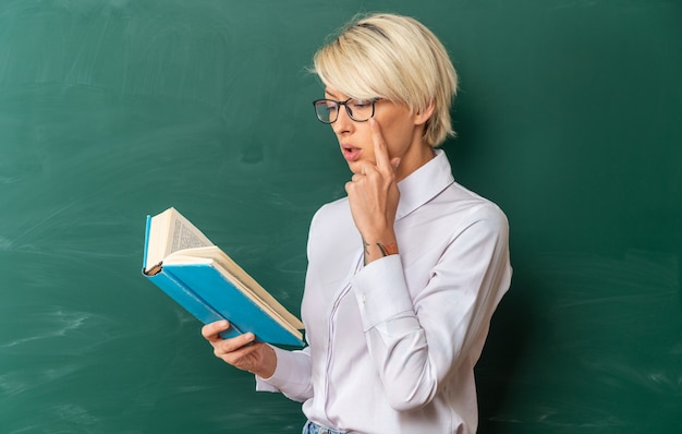 Jeune enseignante blonde concentrée portant des lunettes en classe debout en vue de profil devant le tableau tenant et lisant un livre en gardant la main sur le menton