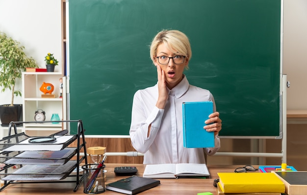 Jeune enseignante blonde concernée portant des lunettes assis au bureau avec des fournitures scolaires en classe montrant un livre fermé gardant la main sur le visage regardant à l'avant