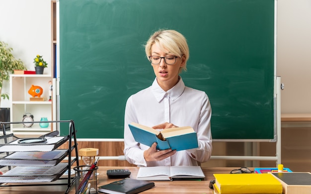 Jeune enseignante blonde impressionnée portant des lunettes assise au bureau avec des outils scolaires en classe tenant et pointant le doigt sur le livre en le lisant