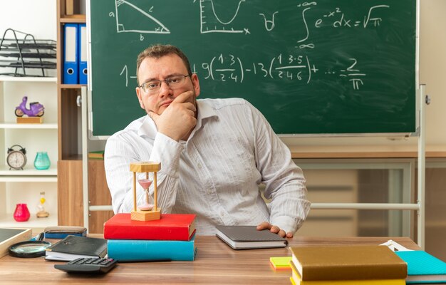 Jeune enseignante blonde réfléchie portant des lunettes assis au bureau avec des outils scolaires en classe en gardant la main sur le menton en regardant la caméra